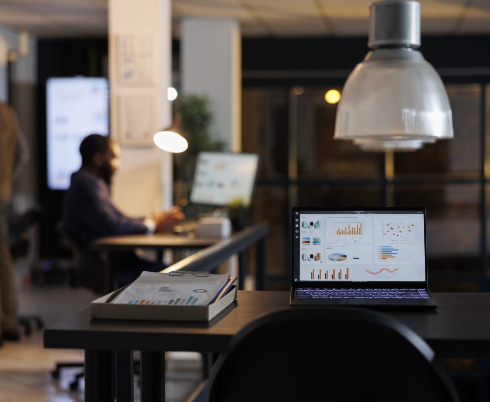 Laptop computer with marketing statistics standing on desk table in startup office, businessmen working overtime at company strategy. African american entrepreneurs brainstorming business ideas